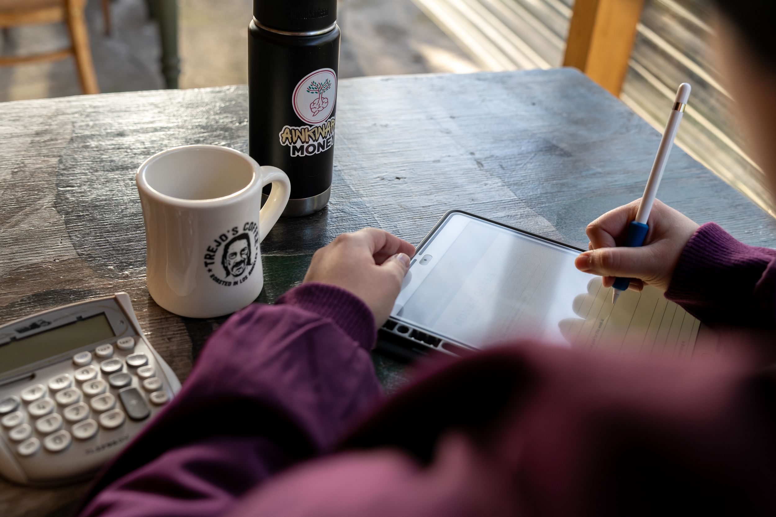 Person writing in notebook at desk with coffee cup and water bottle beside laptop keyboard.