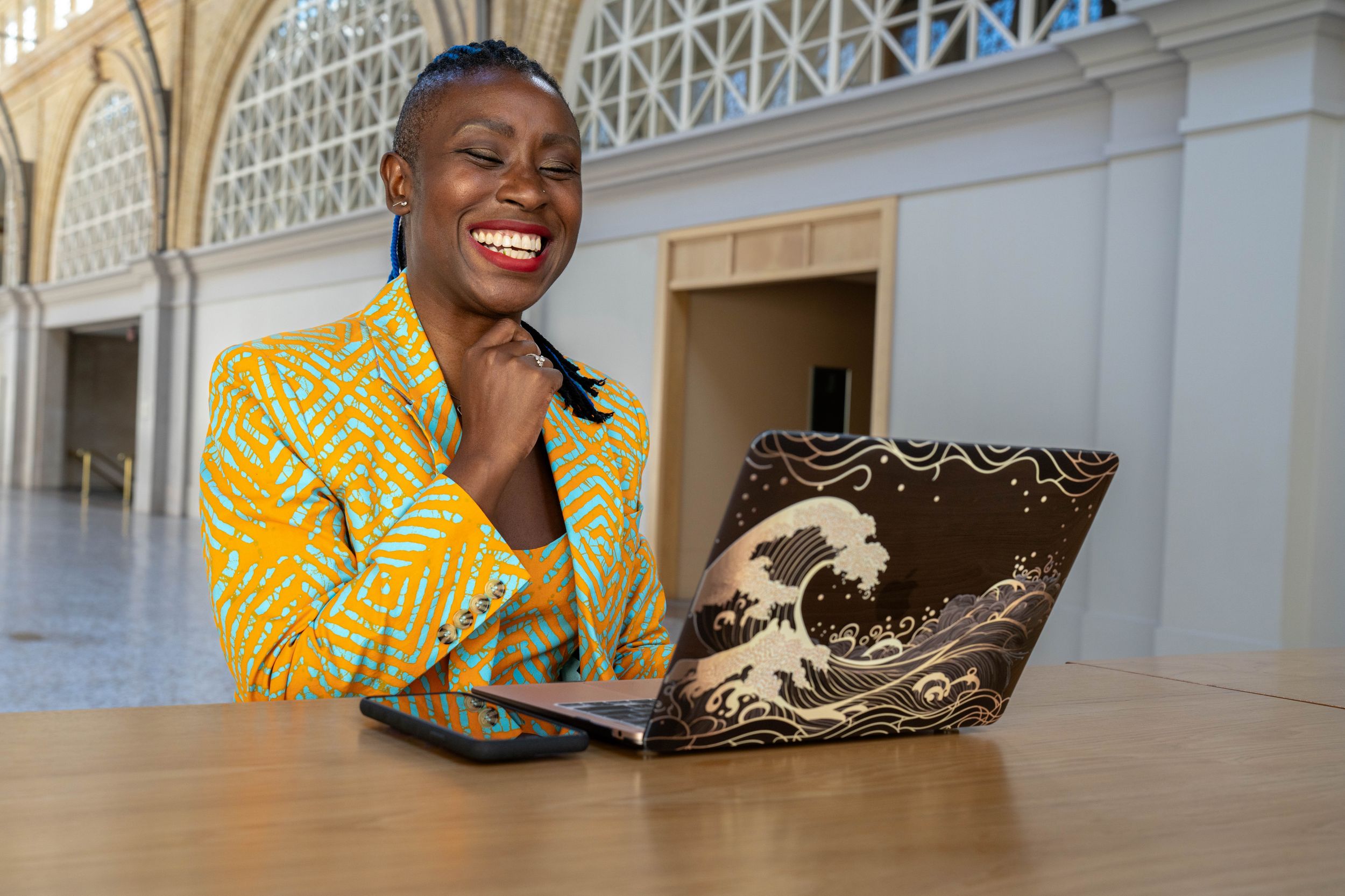 Someone wearing a bright yellow patterned blazer smiles while working on a laptop decorated with a wave design.