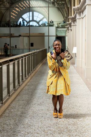 A person in a bright yellow patterned dress and matching shoes shows excitement while standing in an ornate hallway.