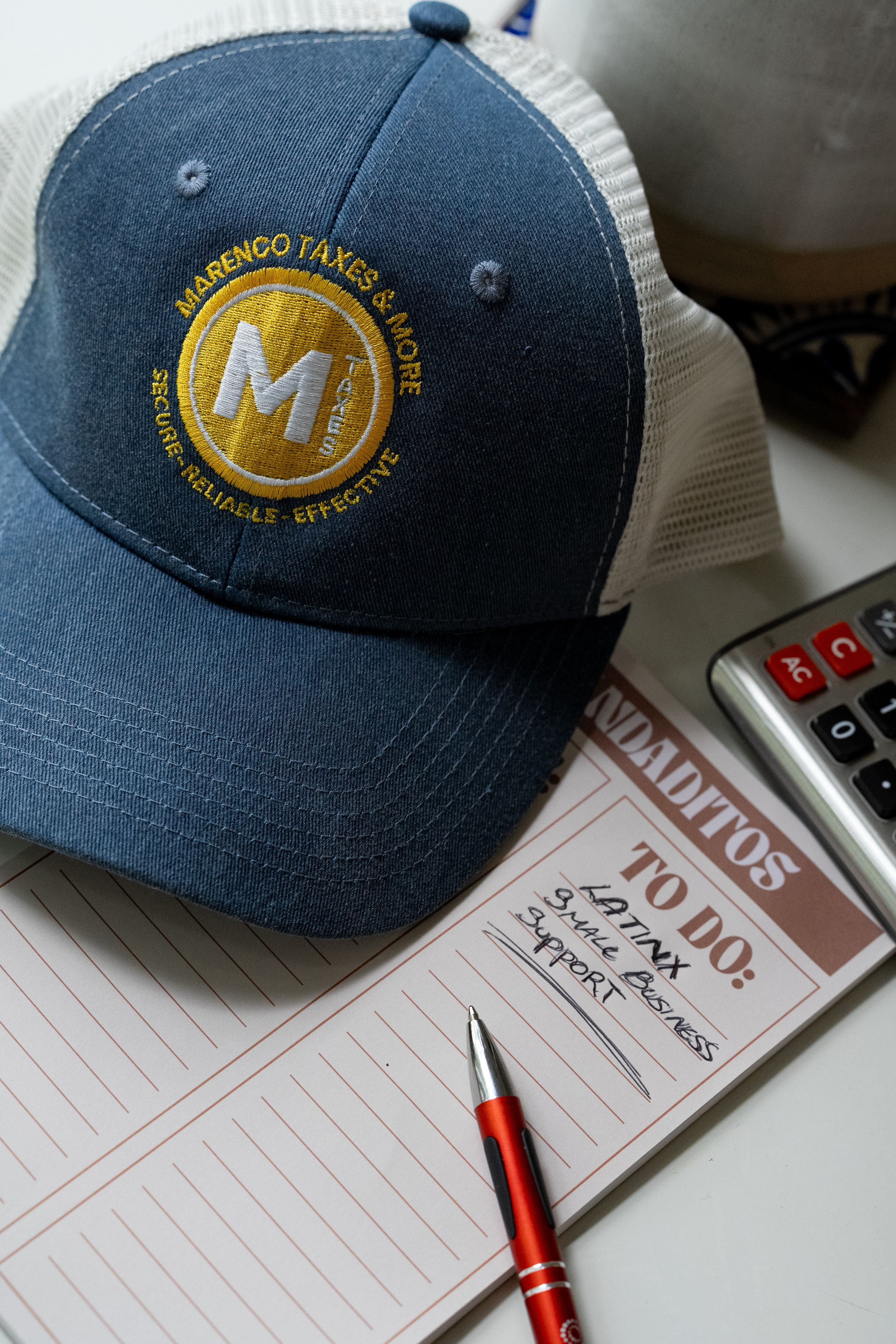 A blue trucker hat with MM logo emblem sits next to a notepad and red pen on a desk with a calculator nearby.