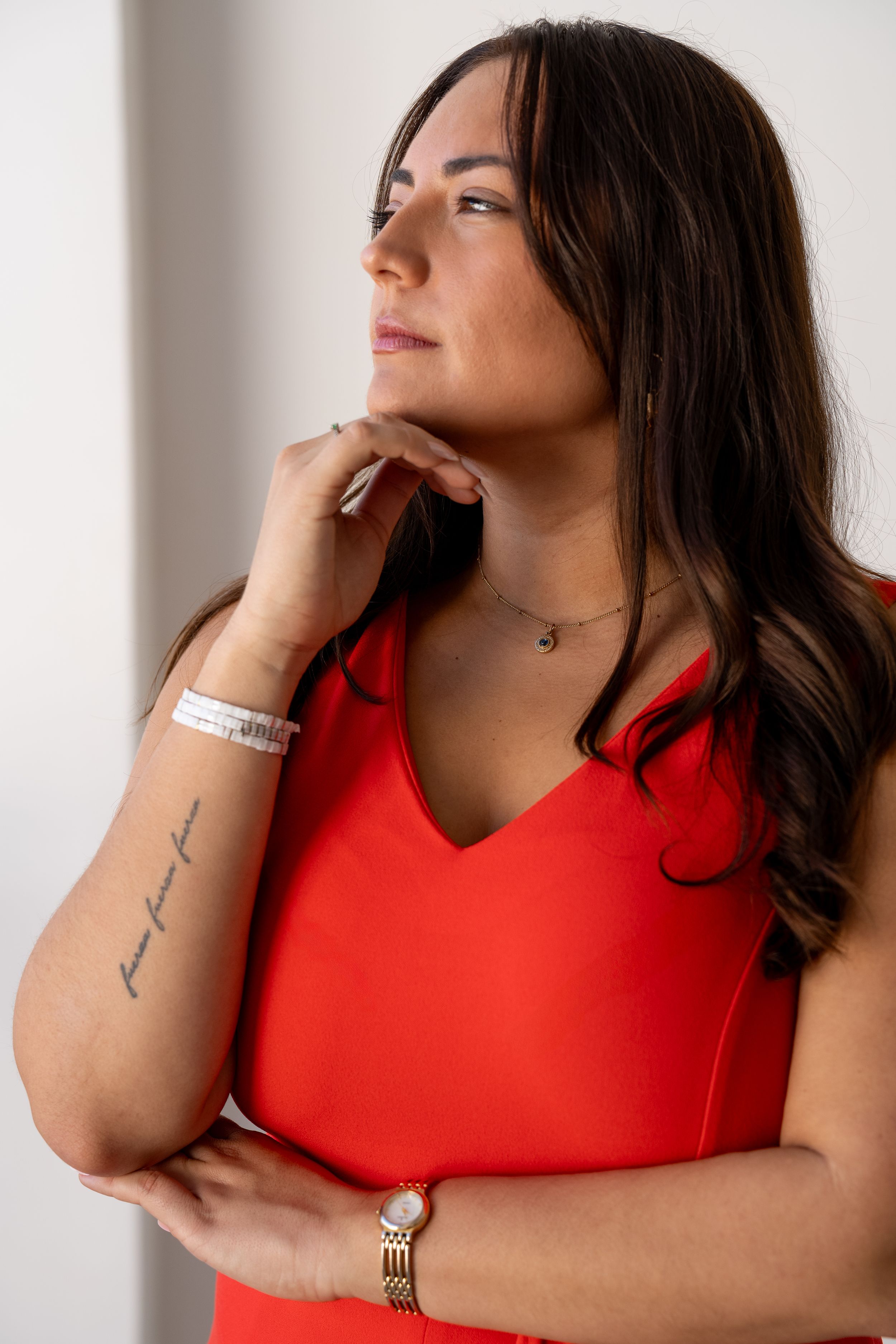 Person in red sleeveless top poses thoughtfully with hand under chin while wearing jewelry and displaying arm tattoo.