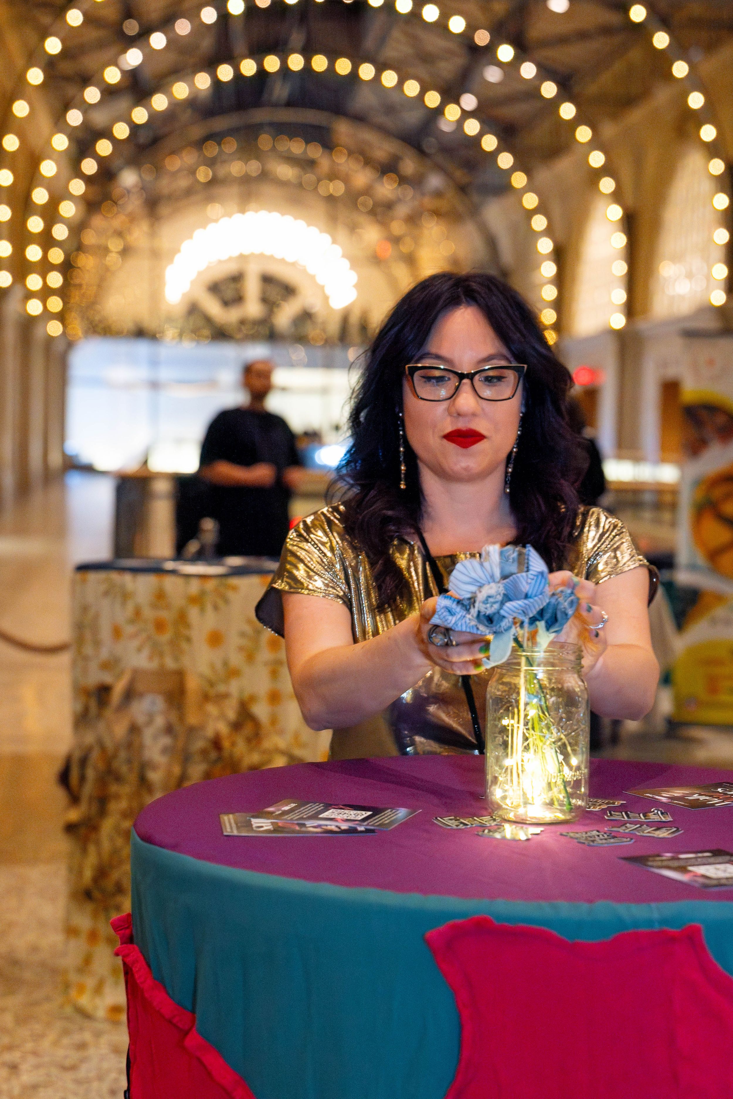 A person in glasses and sparkly top arranges a glowing mason jar on a teal tablecloth under elegant string lights.