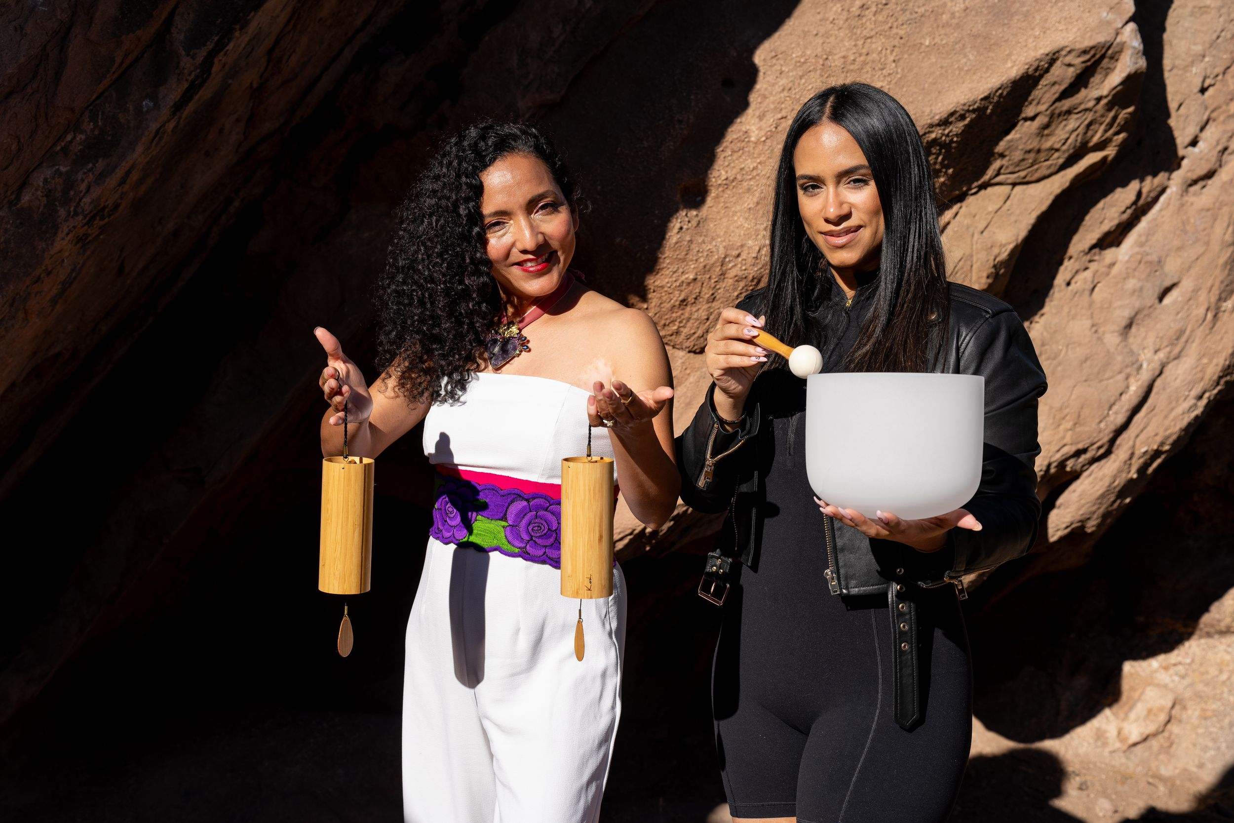 Two people in contrasting outfits hold cooking ingredients while posing against a rocky backdrop outdoors in sunlight.