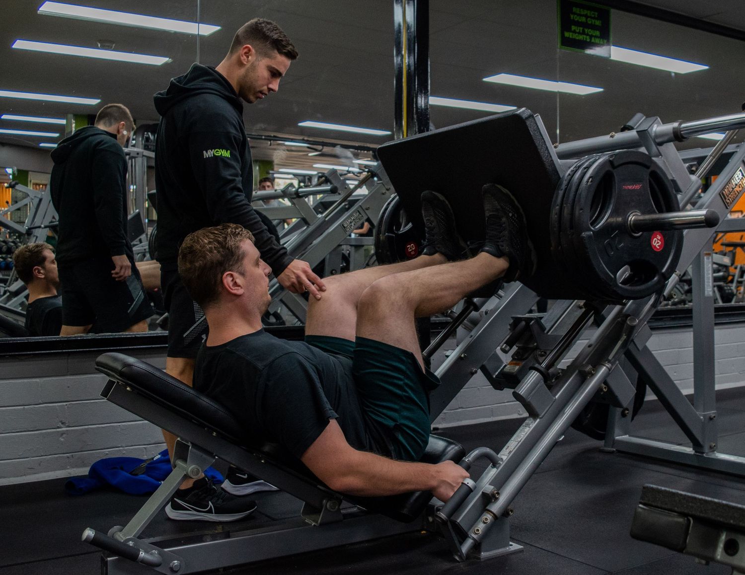 Personal trainer supervising client during leg press exercise at gym.