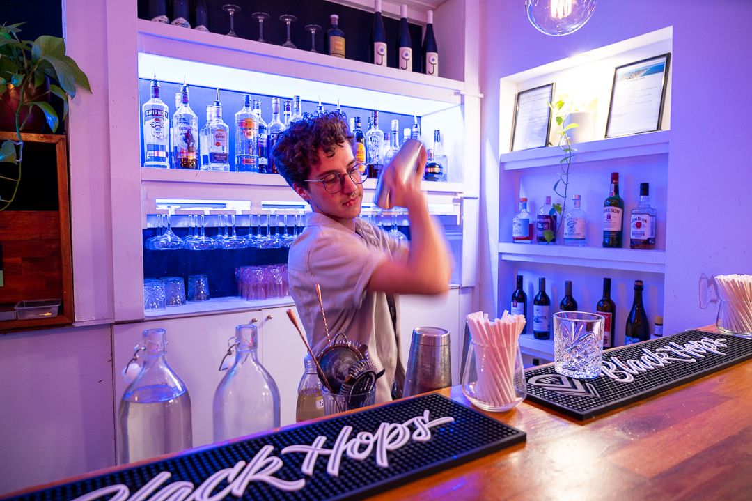 Bartender mixing drinks at illuminated bar counter with purple ambient lighting.