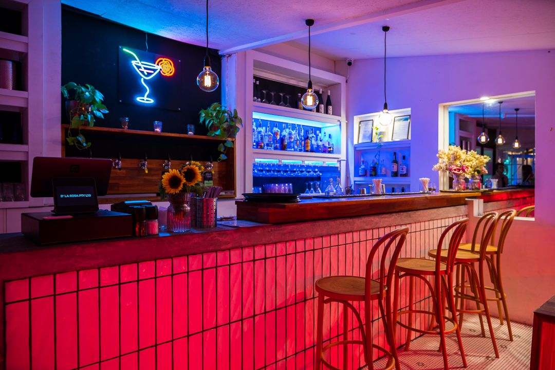 Modern bar interior with red-lit counter, neon cocktail signs, and hanging pendant lights.