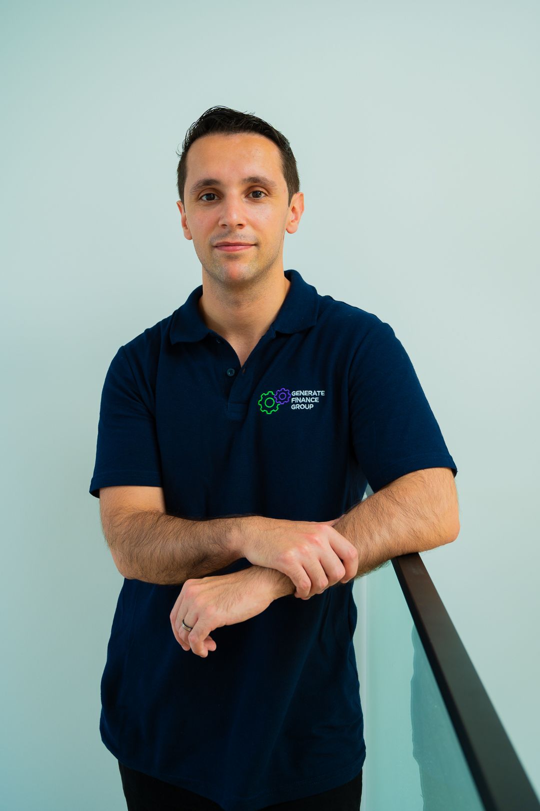 Business owner in dark blue polo shirt poses casually against light colored wall.