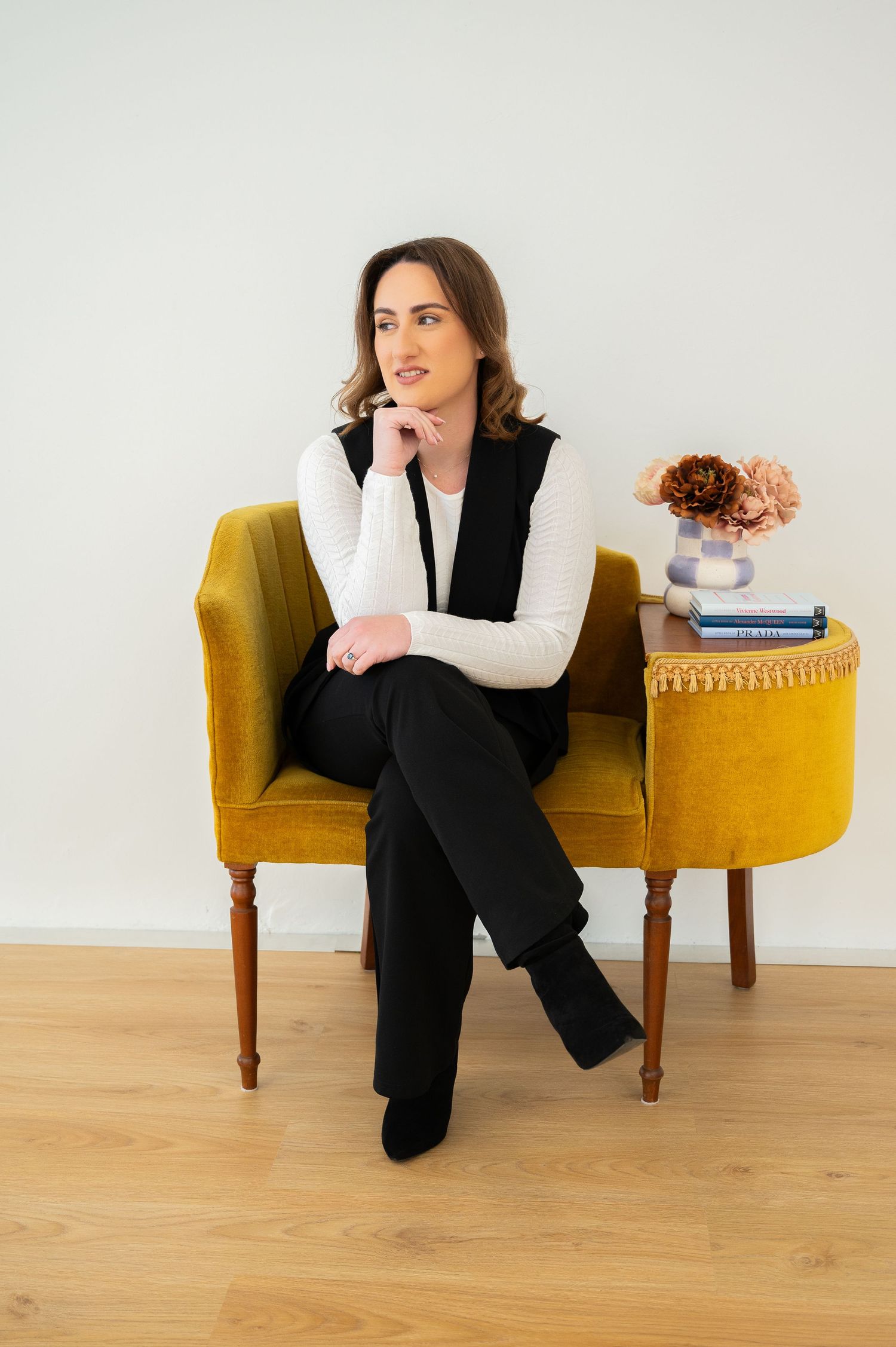 Person in black business attire sits in various poses on a mustard yellow bench against white wall.