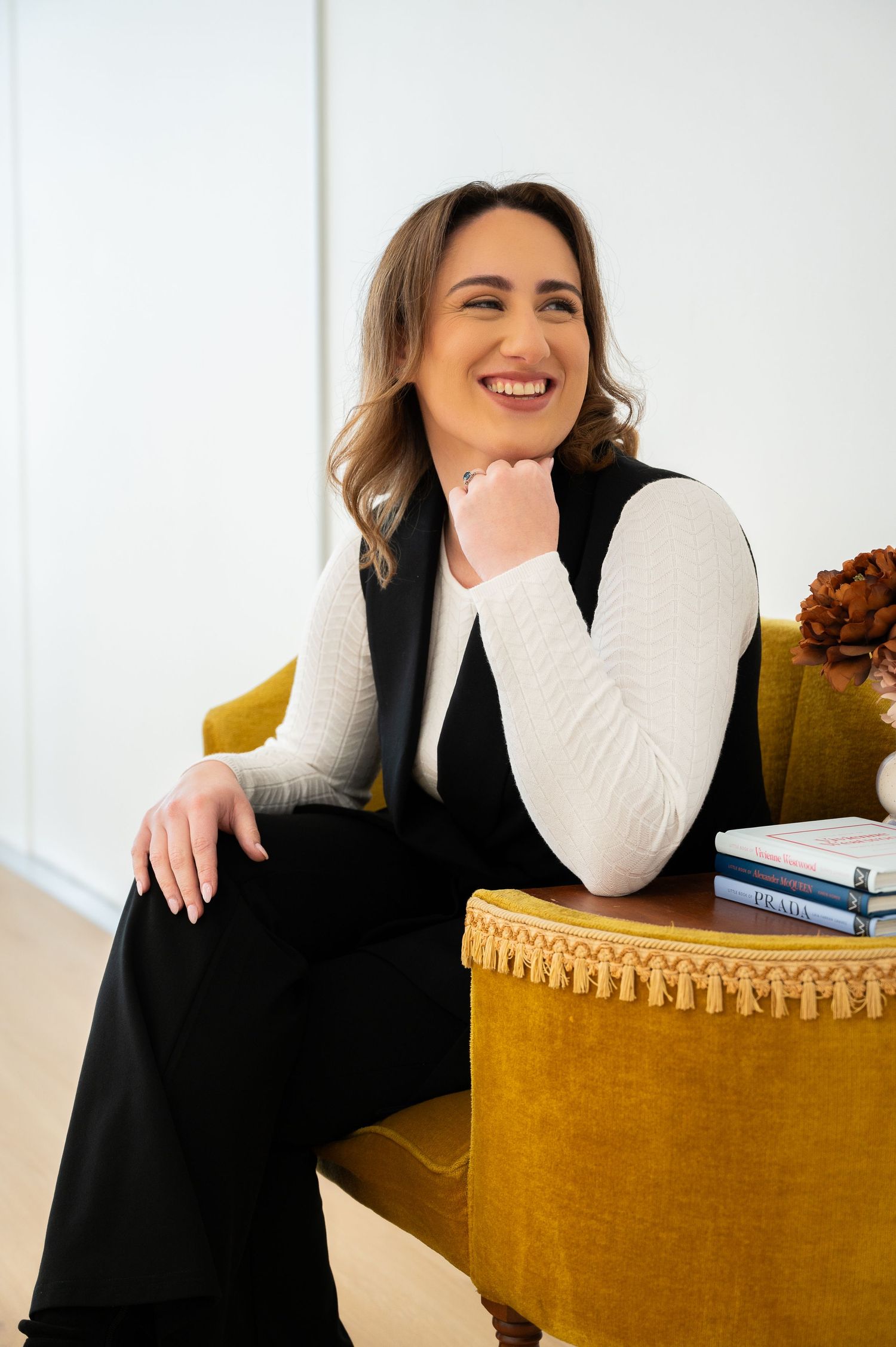 Professional wearing black blazer and white sweater laughs joyfully while posing on gold velvet couch in bright room.