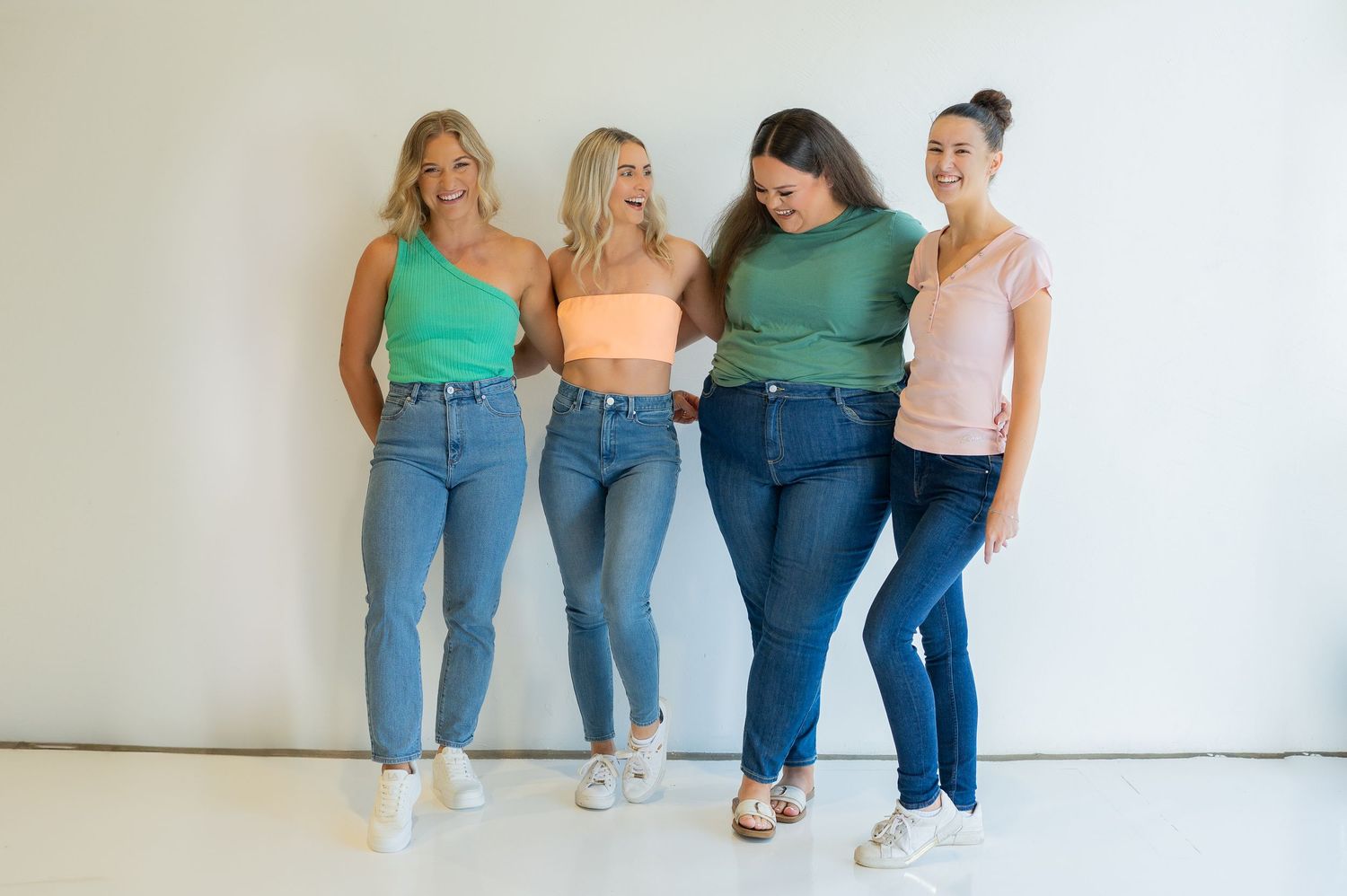 Four people lean against a white wall wearing blue jeans and different colored tops in a casual group photo.