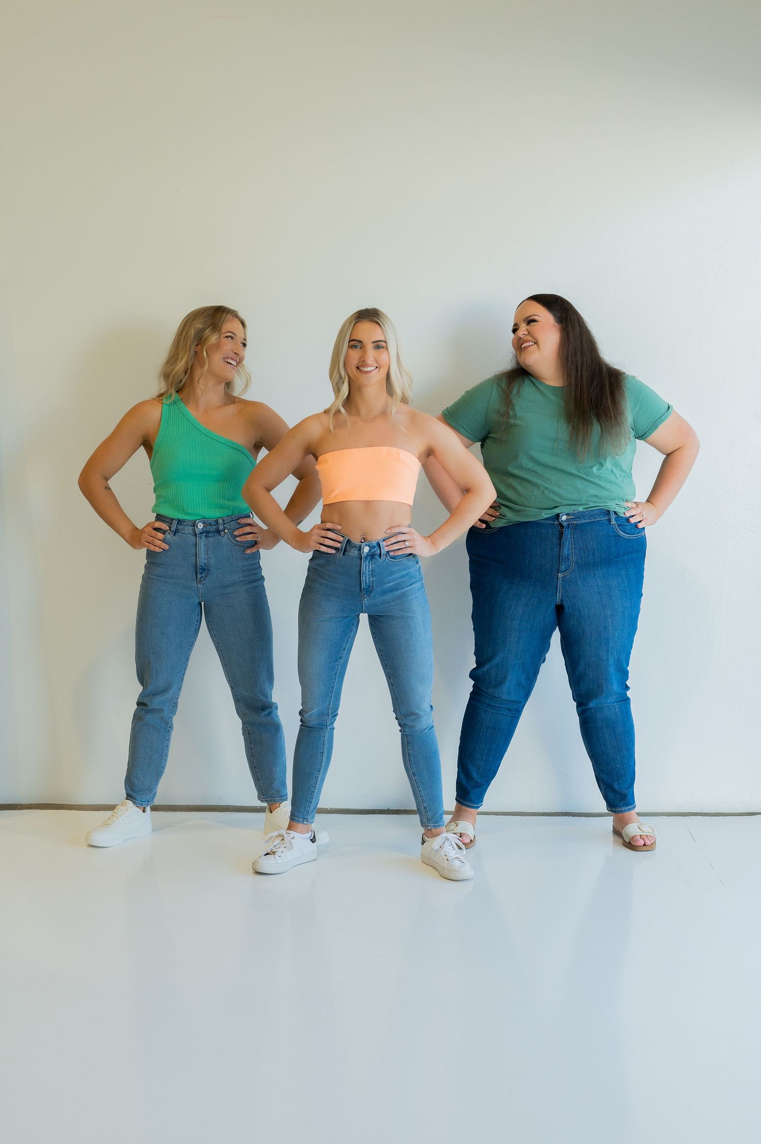Three models strike poses showing off different styles of blue jeans and crop tops against a white wall.