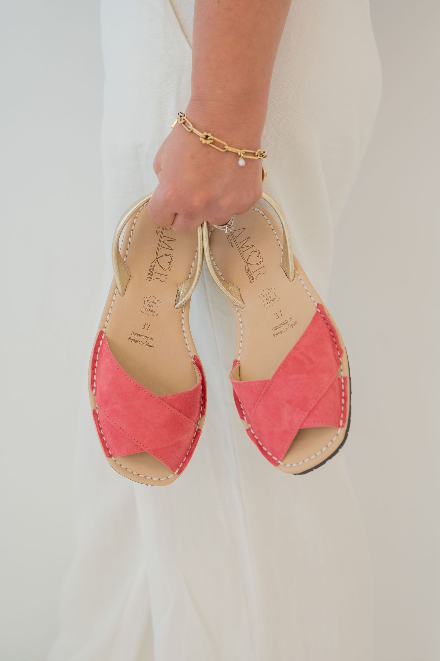Coral pink suede peep-toe sandals with gold ankle bracelet shown against white background.