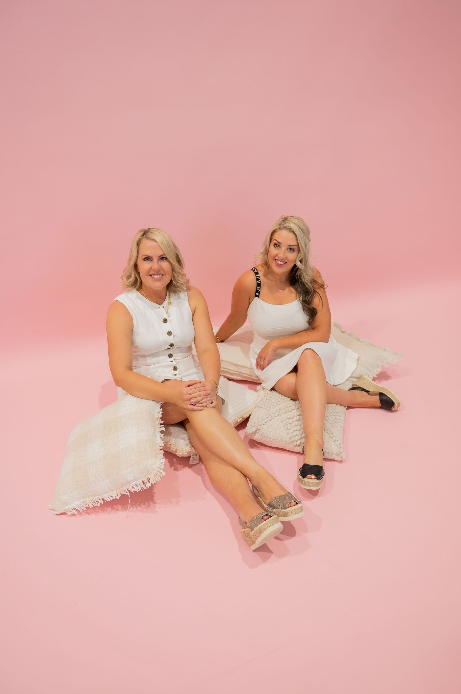 Two women in white dresses sit together on cushions against a pink studio backdrop.
