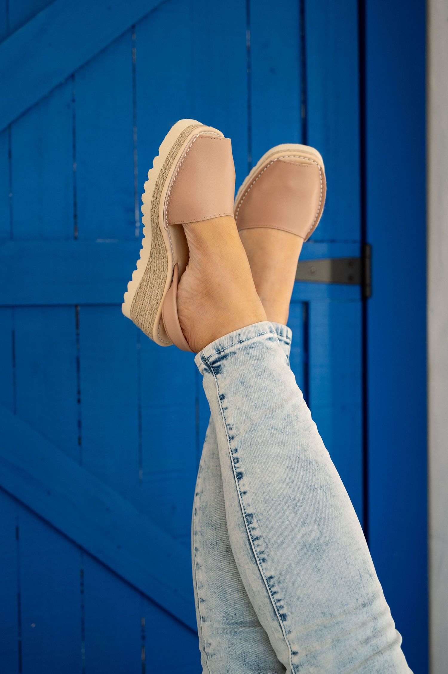 Legs wearing light acid wash jeans and beige platform loafers raised up against a bright blue wooden door.