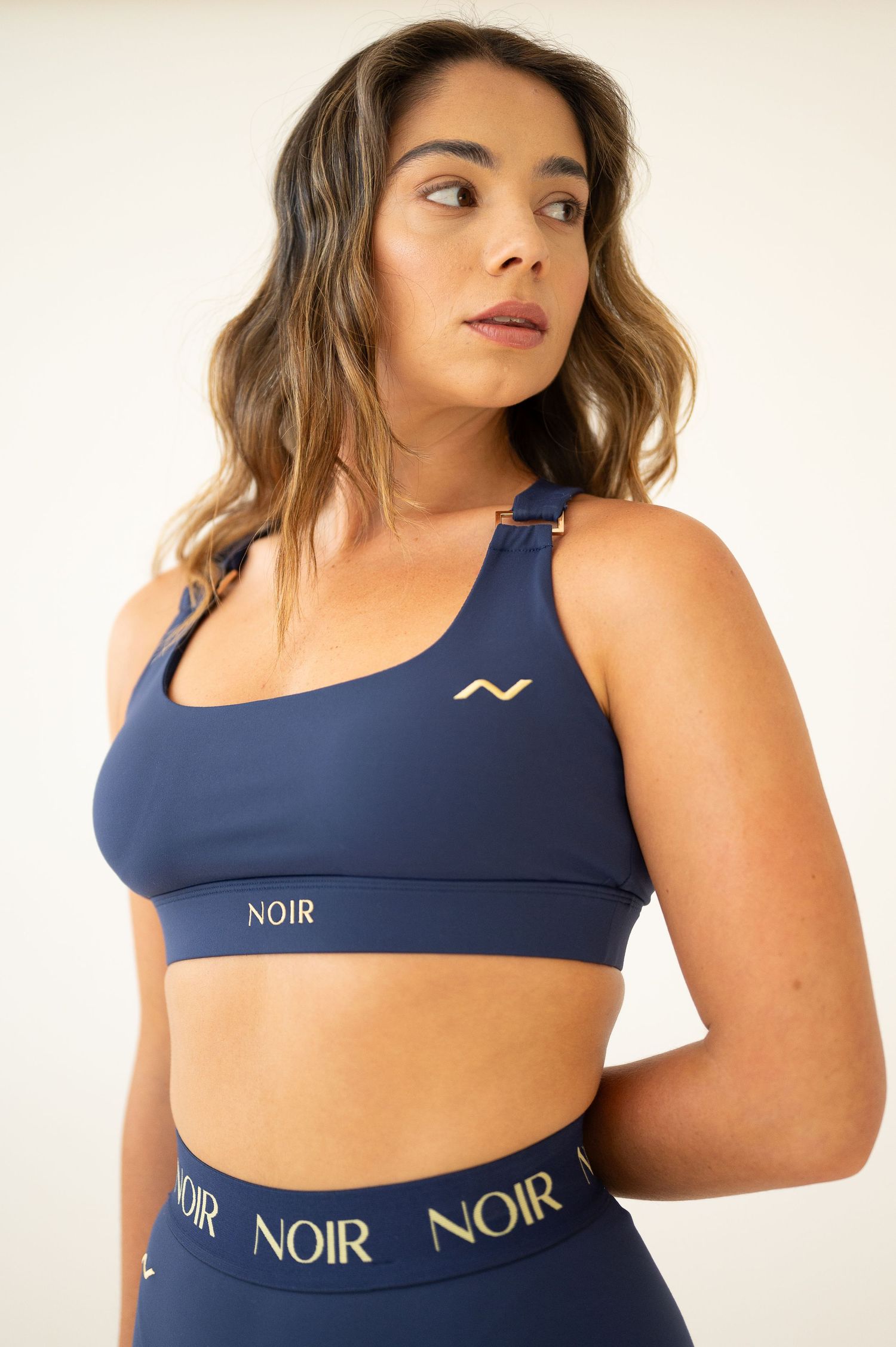 Athletic model wearing navy blue sports bra and leggings with Noir branding poses against light background.