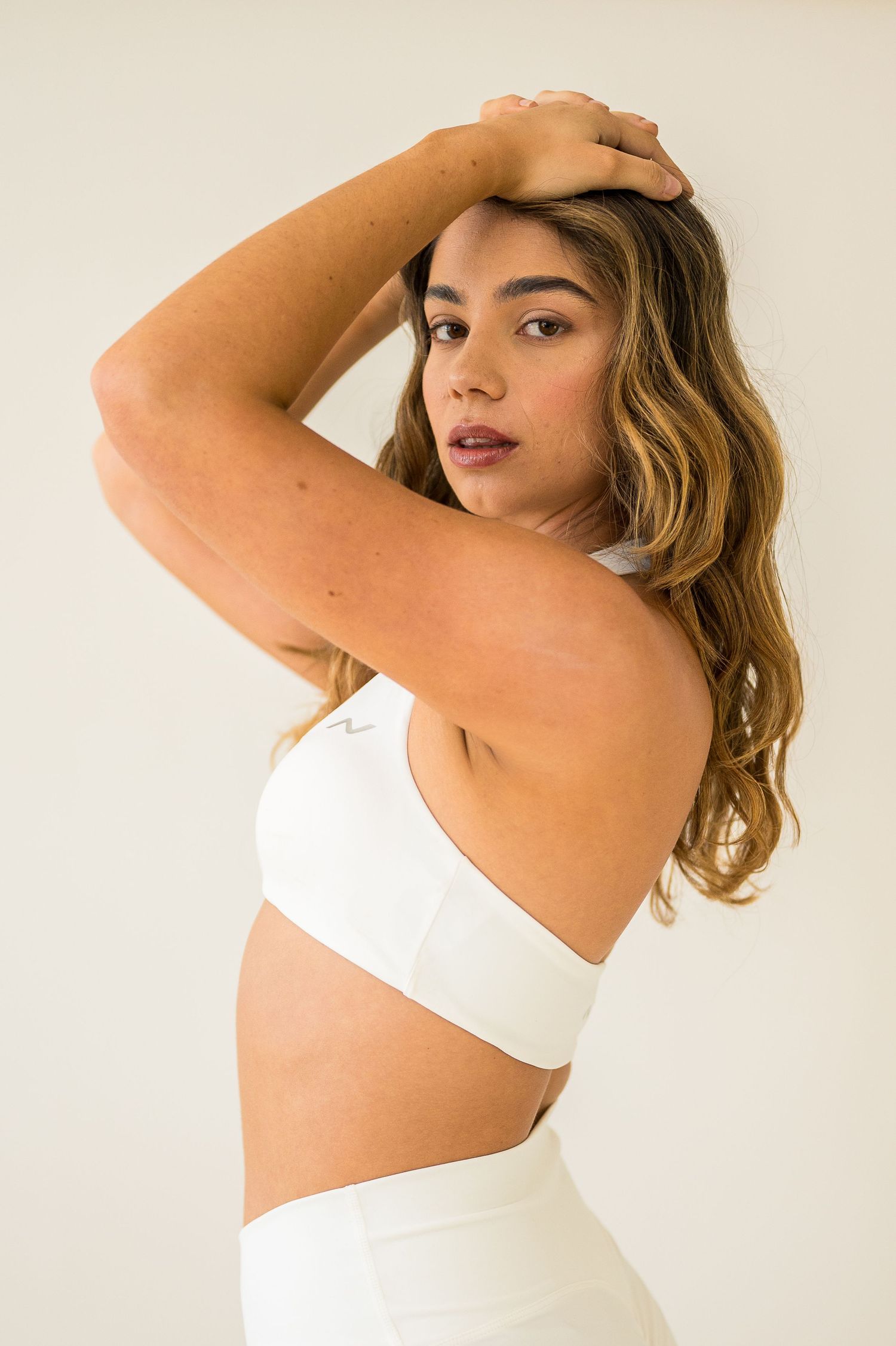 Fashion model in white crop top and high-waisted bottoms poses against neutral background with hand in wavy hair.