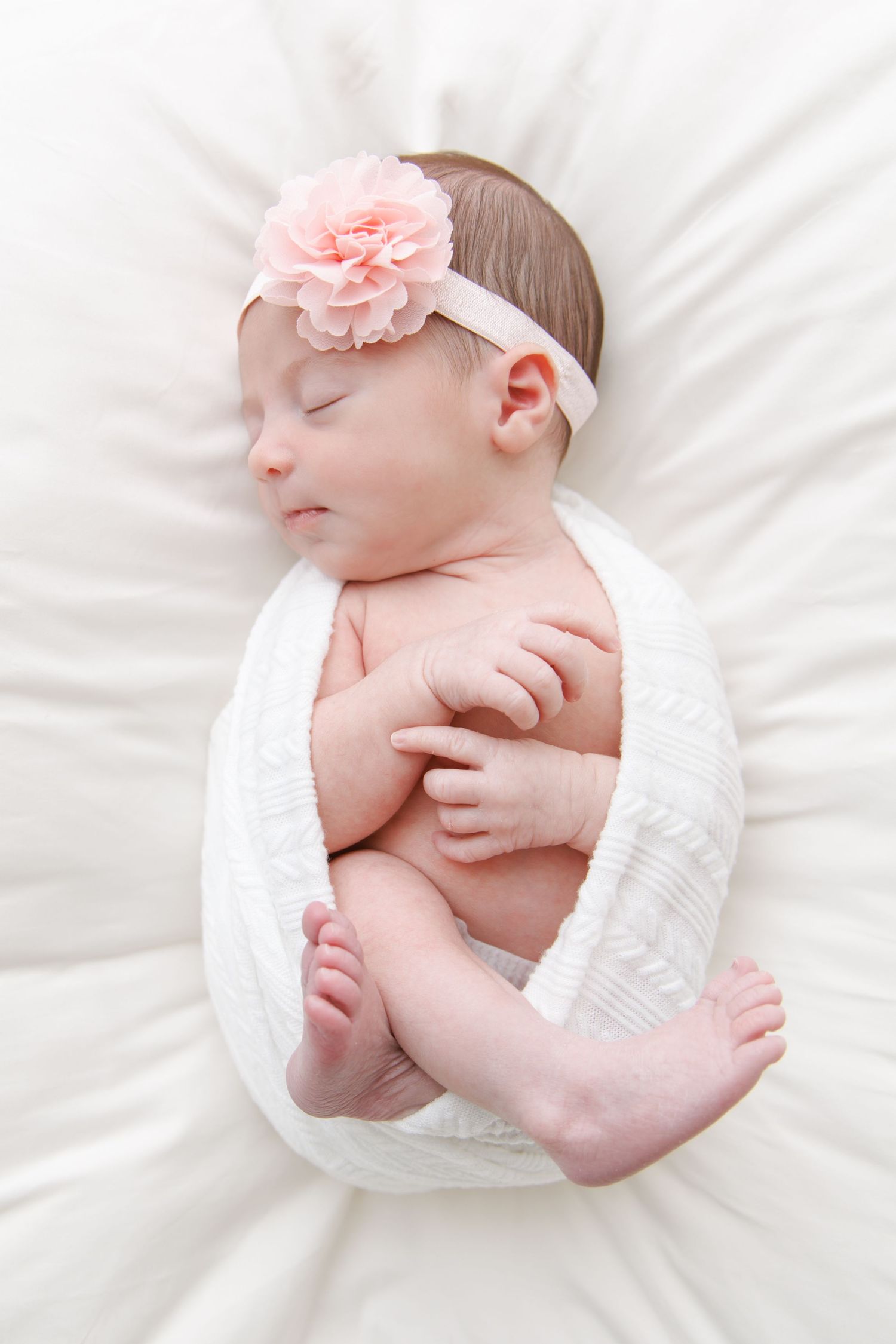 A newborn baby wearing a diaper rests peacefully on white bedding with a pink flower headband.