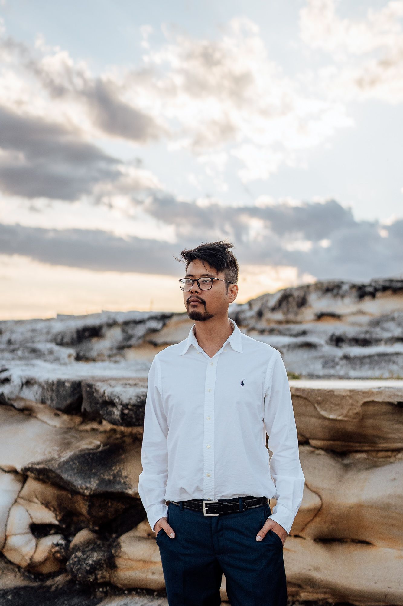 Person in white shirt and dark pants poses against dramatic rocky coastal landscape at sunset.