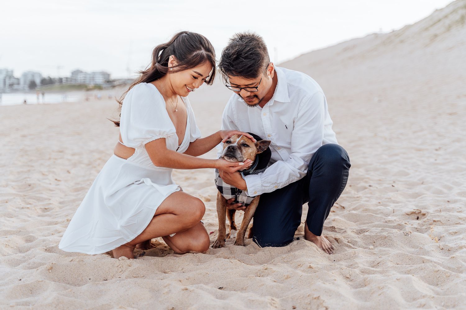 Couple and dog share tender moments on a sandy beach during a sunset photoshoot.