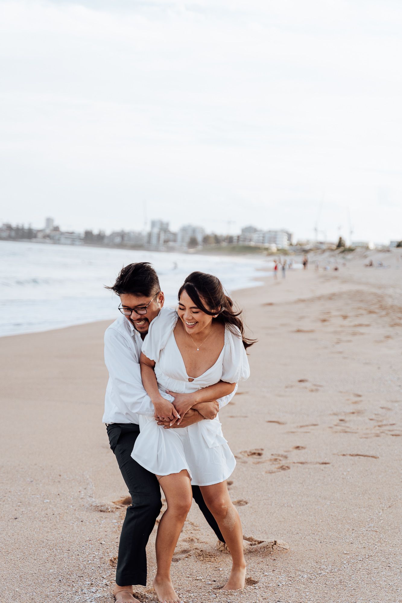 A couple playfully dances together on a sandy beach with a cityscape visible in the background.