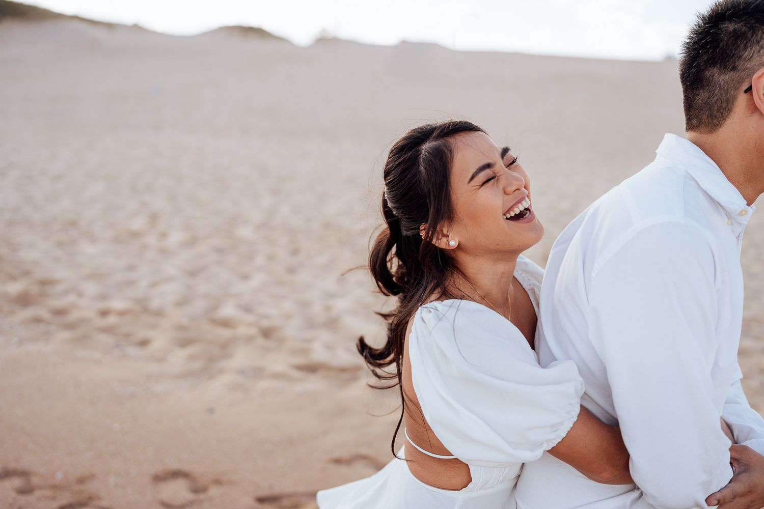 A couple in white outfits shares romantic moments on a sandy beach during a sunset photoshoot.