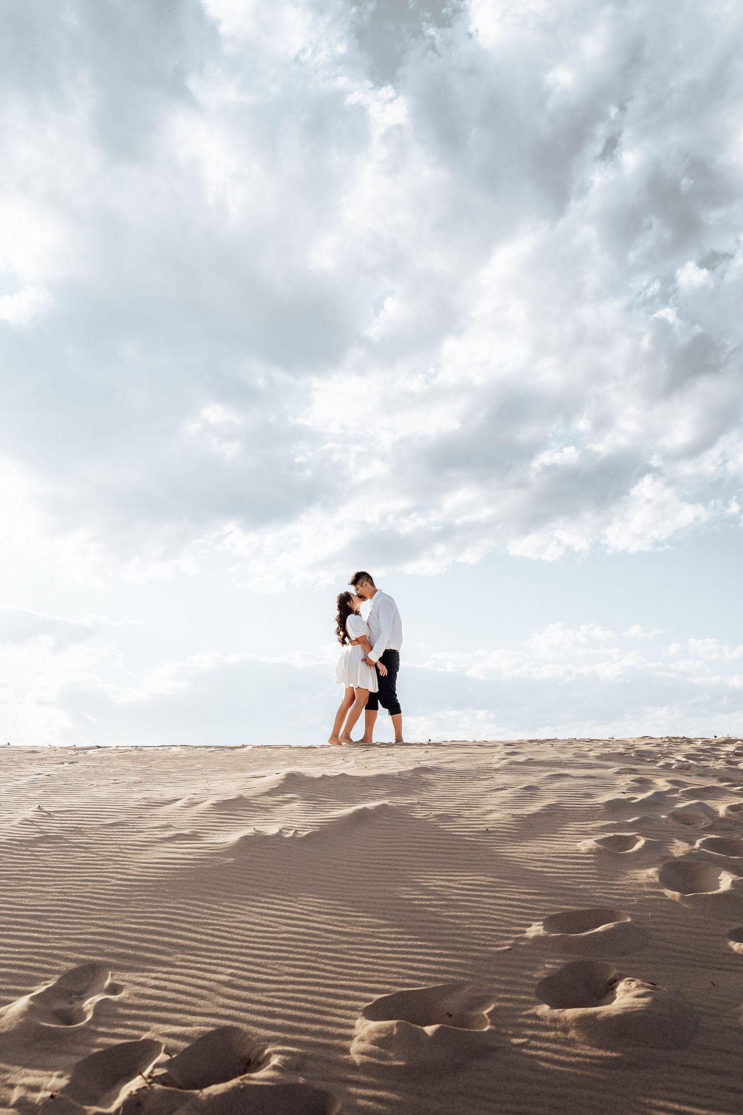 A couple embraces on a sandy dune under dramatic cloudy skies at the beach.