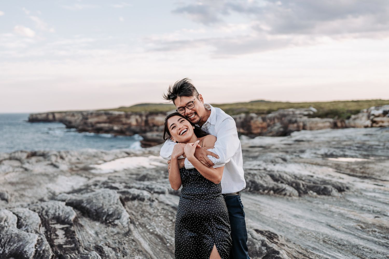 A couple embraces on rocky coastal cliffs at sunset with ocean views in the background.