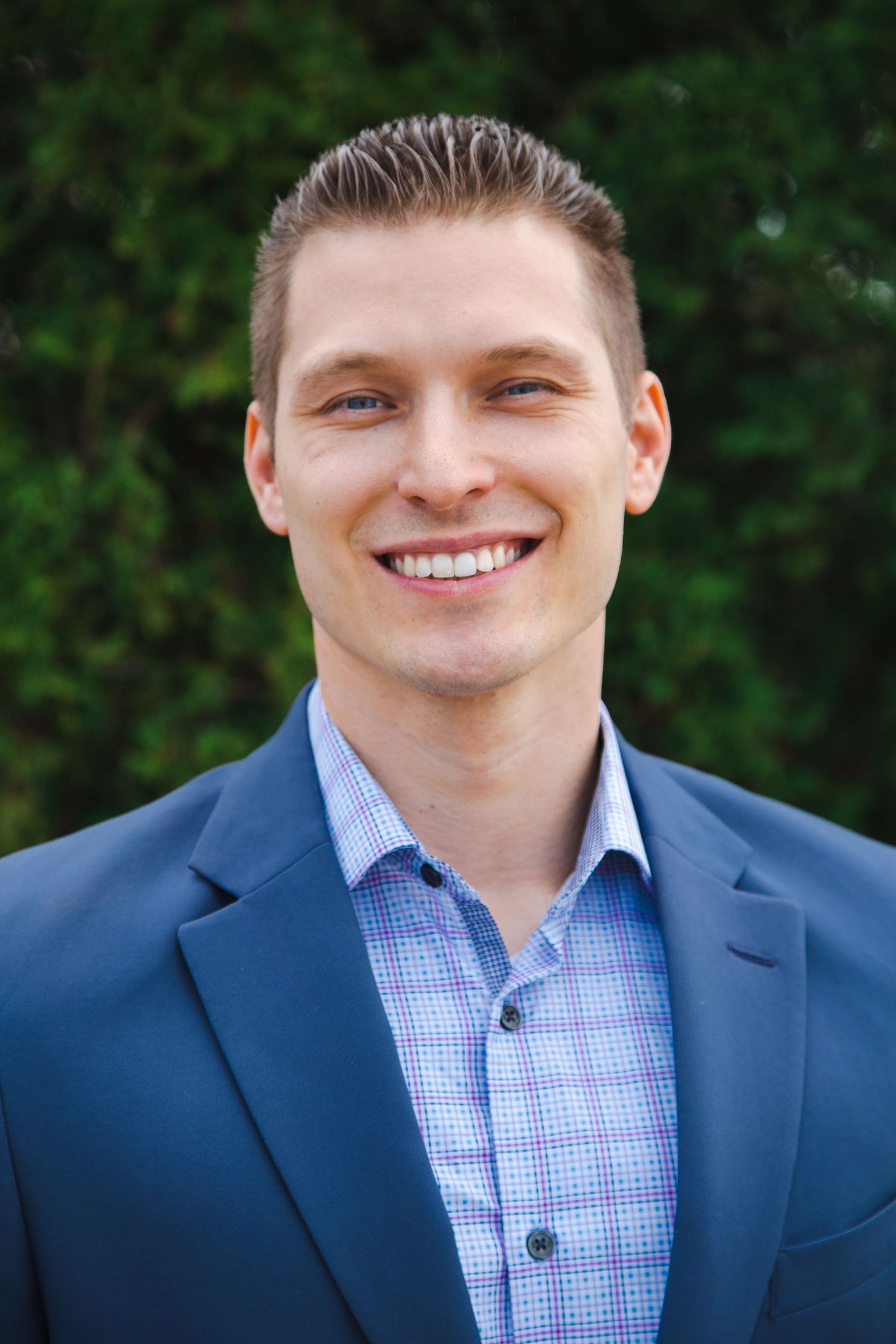 Professional headshots of someone in a navy suit and checkered blue dress shirt against a green outdoor background.