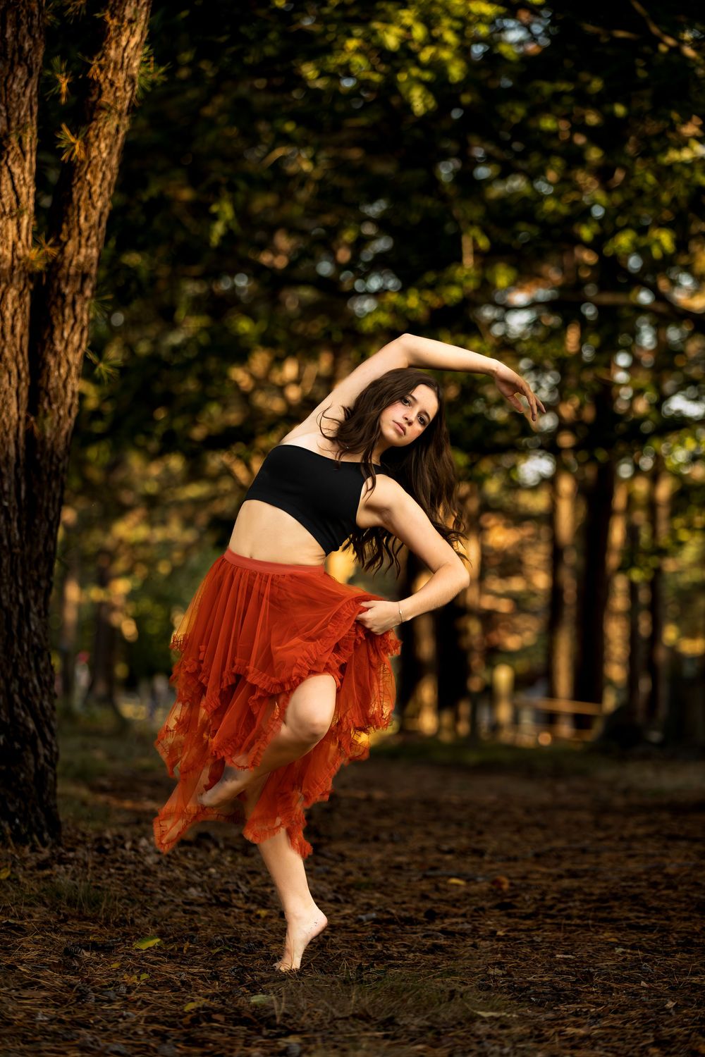 Dancer in flowing red tulle skirt performs graceful movements in sunlit autumn forest setting.