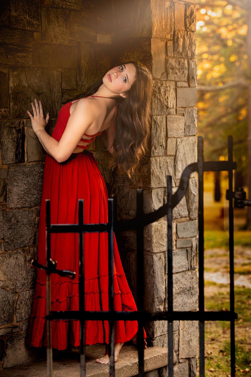A person in a vibrant red dress poses dramatically against a stone wall with iron bars during autumn.