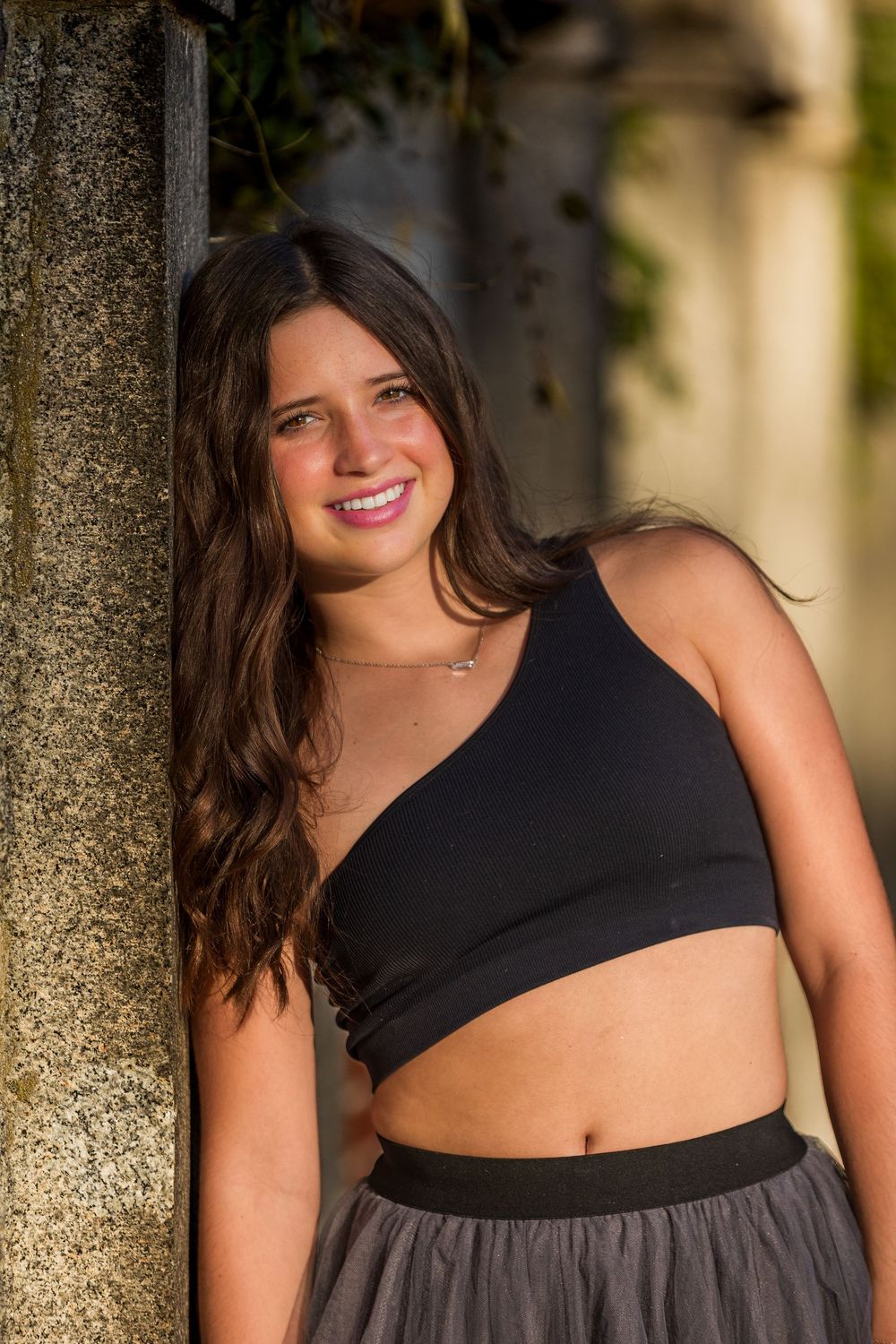A person wearing a black crop top and gray skirt poses outdoors in warm sunlight against a stone pillar.