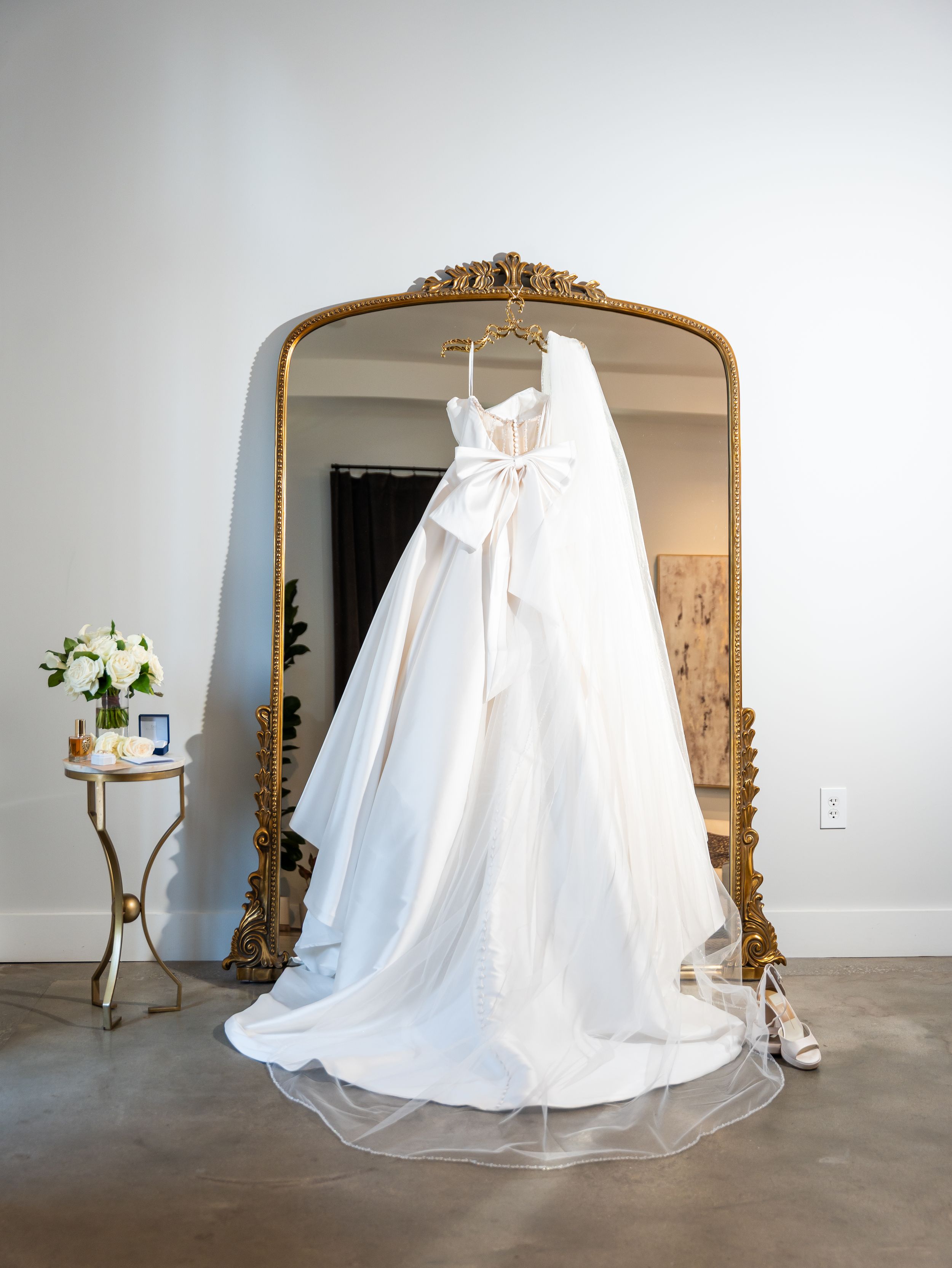 Elegant white wedding dress hanging on ornate gold mirror with floral arrangement on side table in Raleigh NC