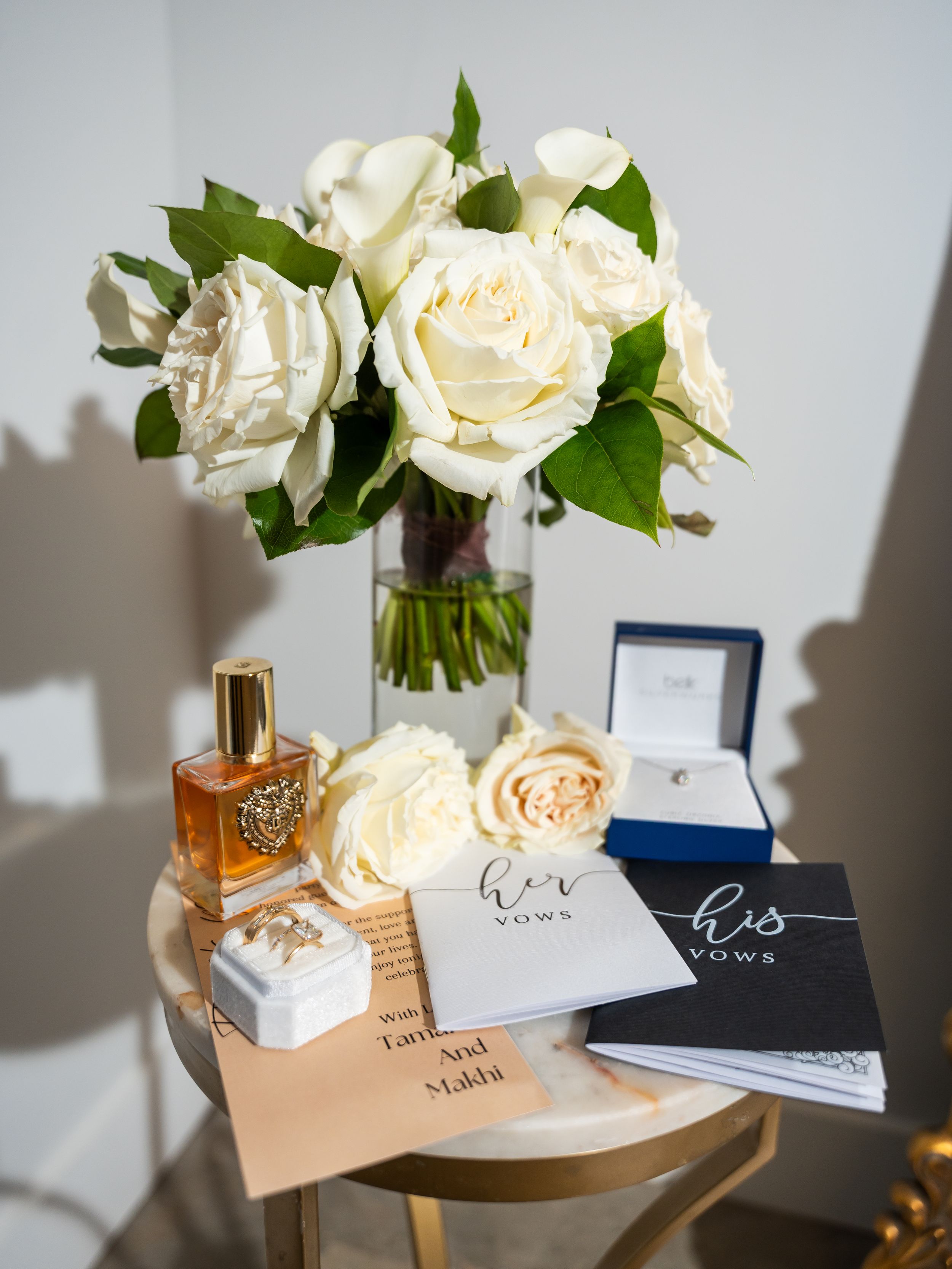 Wedding details with white roses, perfume, and invitation cards on gold accent table.