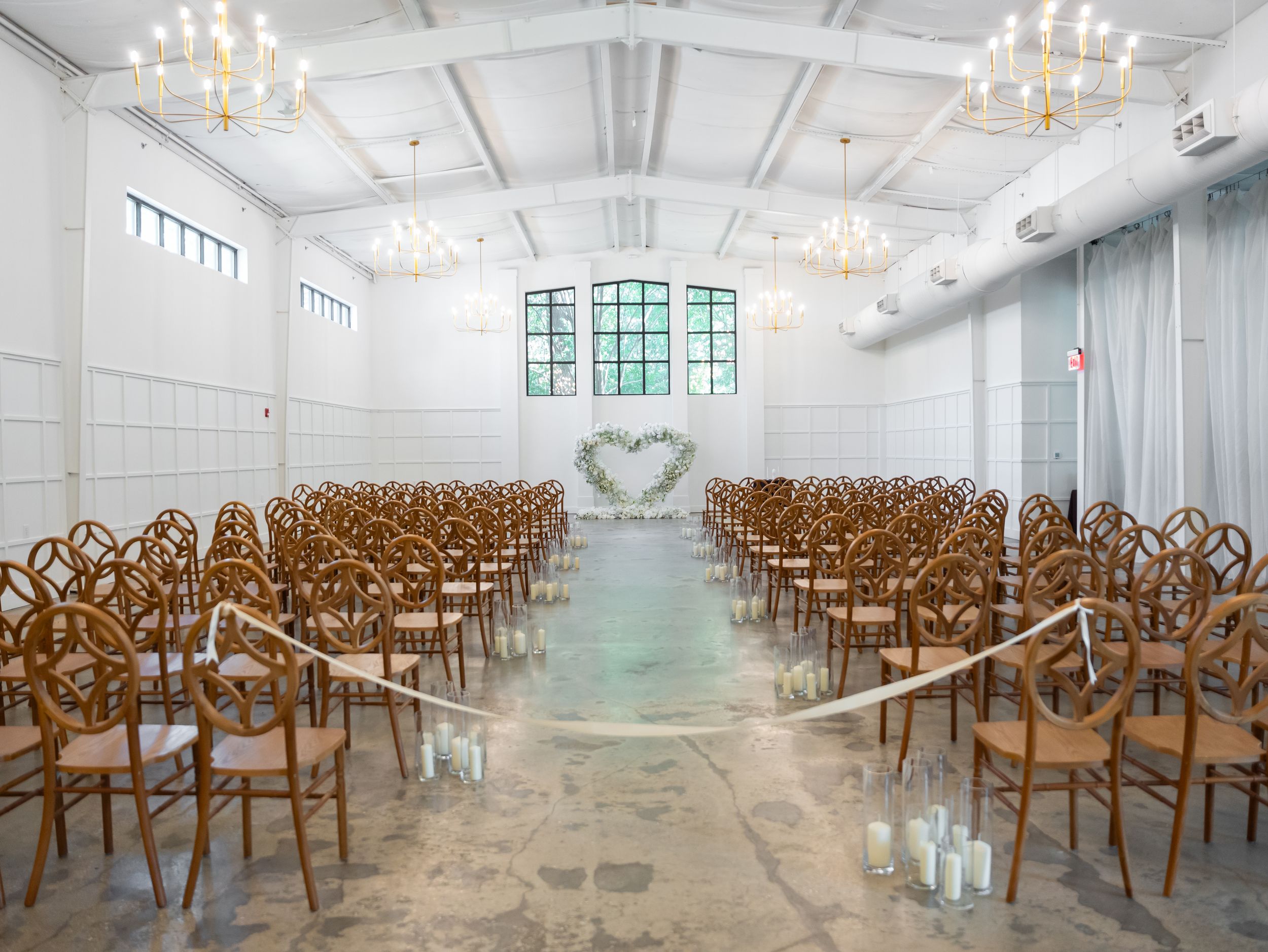 Modern white wedding ceremony venue with wooden chairs arranged in rows and chandeliers overhead.