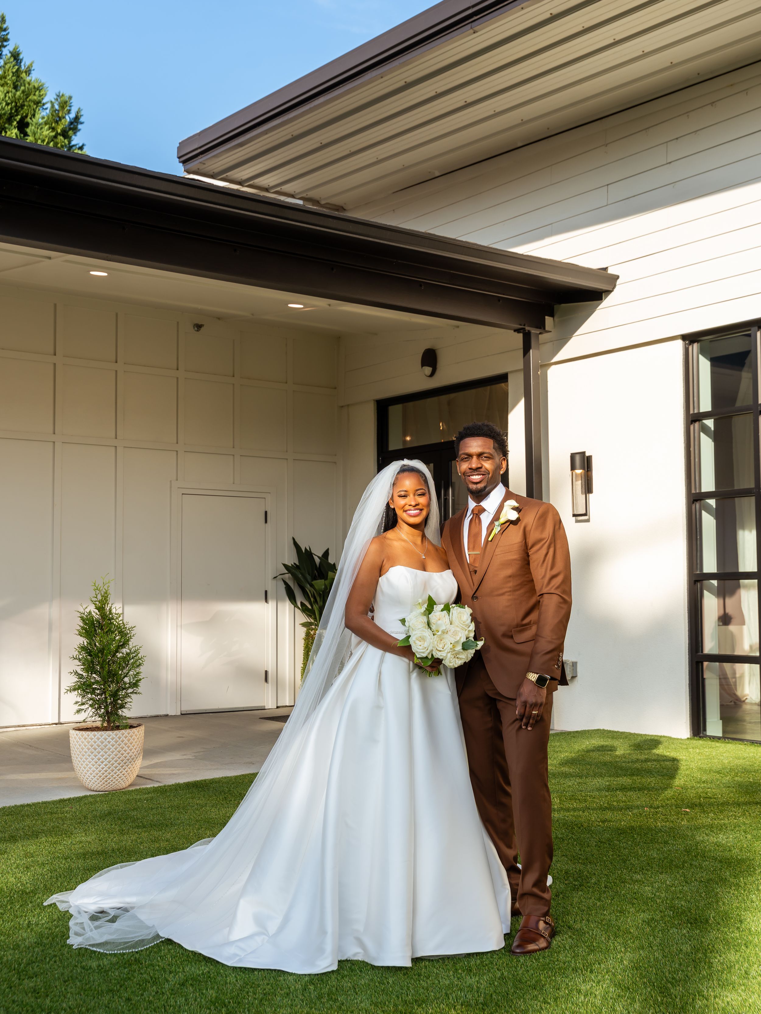 A couple in wedding attire poses together on a grassy lawn outside a modern white building.