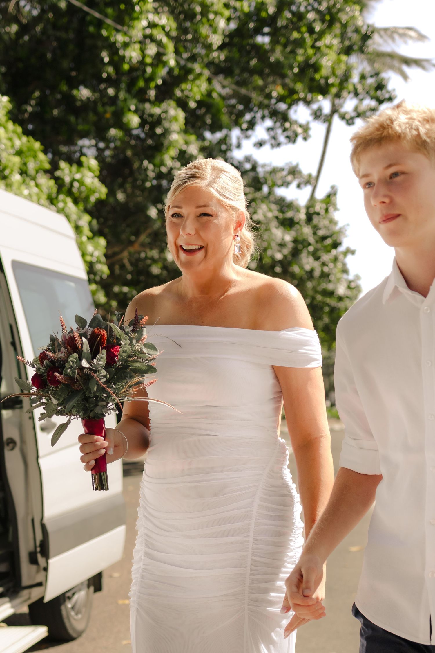 Wedding party in white attire gathering outdoors near a vehicle.