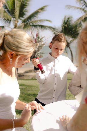A young boy in white holds a red bottle during a beachside wedding ceremony.