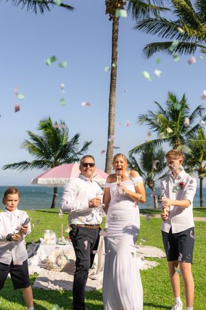 A wedding party celebrates with confetti flying under palm trees by the ocean.