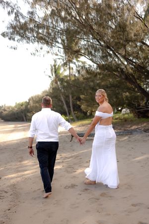 Couple walking hand in hand along a scenic beach with trees providing shade.