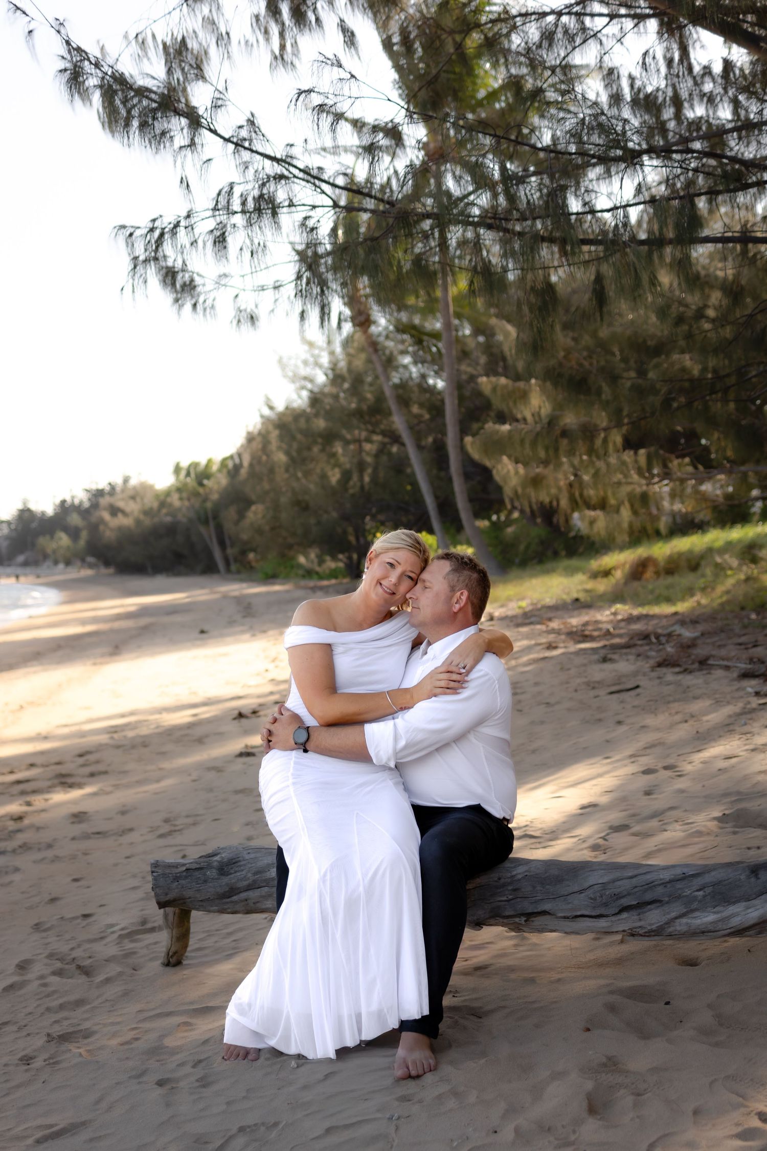 Couple sharing tender moment on beach under overhanging trees.