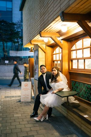 Couple in formal wear sitting on wooden bench under warm lighting.