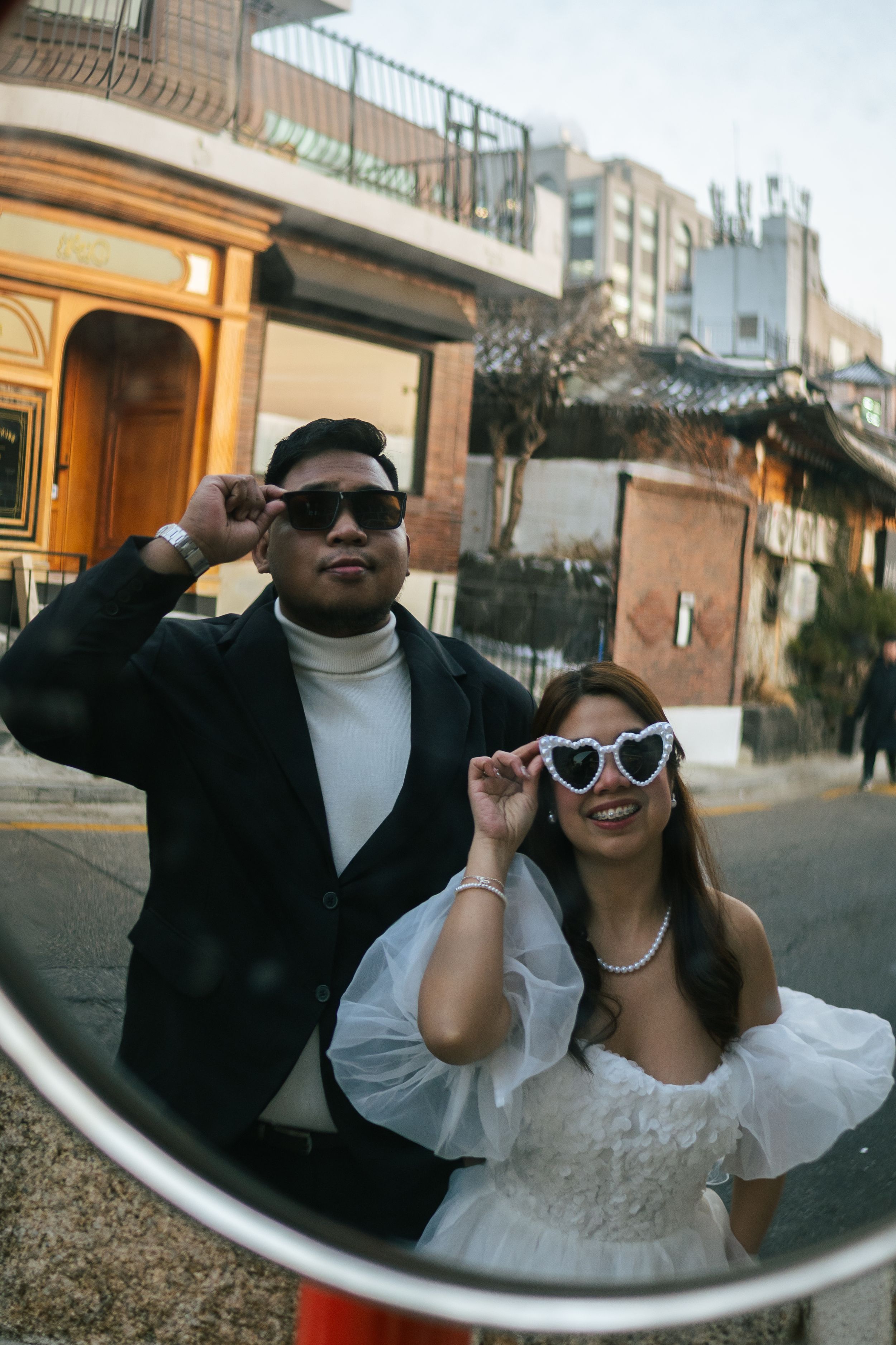 Playful couple posing with sunglasses against urban brick wall background.