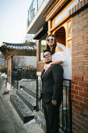 Couple in formal attire standing by brick building entrance with retro sunglasses.