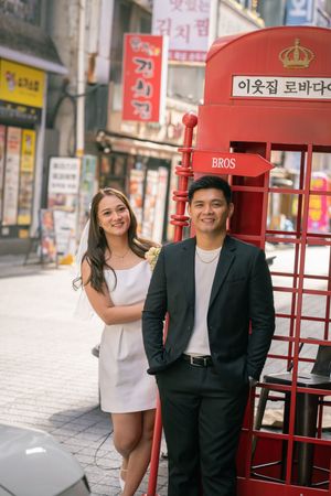 Couple poses near red British-style phone booth with Korean signage in background.