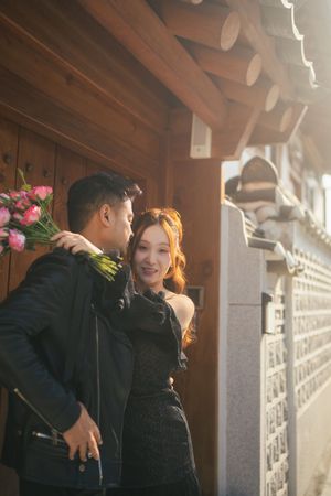 Two people share an intimate moment in a sunlit hallway with wooden beams and white walls.