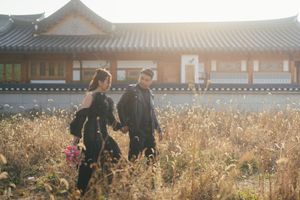 A couple walks through a sunlit field of tall golden grass near traditional Asian architecture.