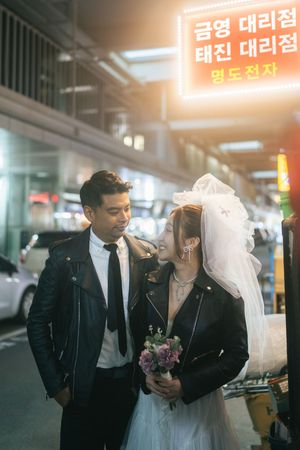 A bride and groom share romantic moments in front of illuminated Korean storefront signs at night.
