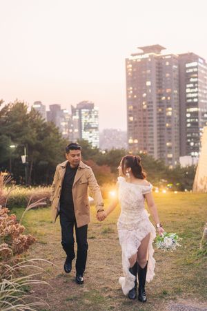 A couple walks together through an urban park at sunset with city buildings in the background.