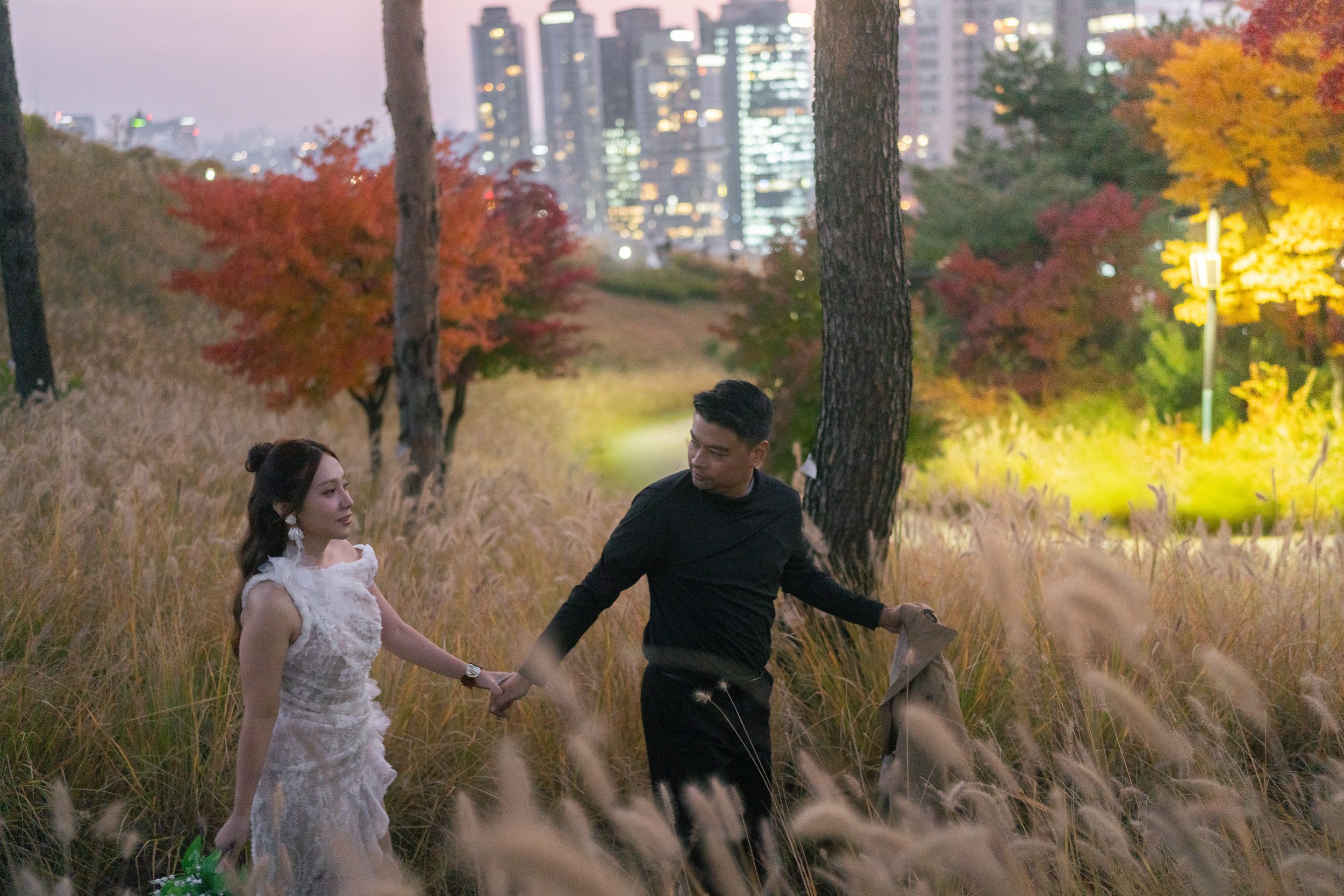 A group of dancers performs outdoors against a backdrop of autumn trees and city lights at dusk.