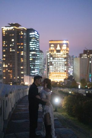 Two people share an intimate moment on a rooftop with illuminated city buildings creating a romantic nighttime backdrop.